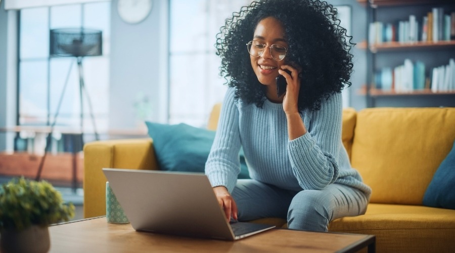 Pretty, black Latina with curly hair and glasses, sitting on yellow sofa, on cell phone while perusing laptop, seeking a structured settlement quote.