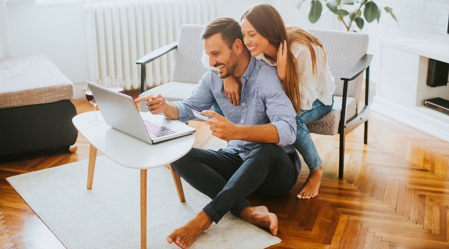 Individuals can buy annuity payments — a couple is smiling, the man sitting on floor and woman right behind him looking at laptop.