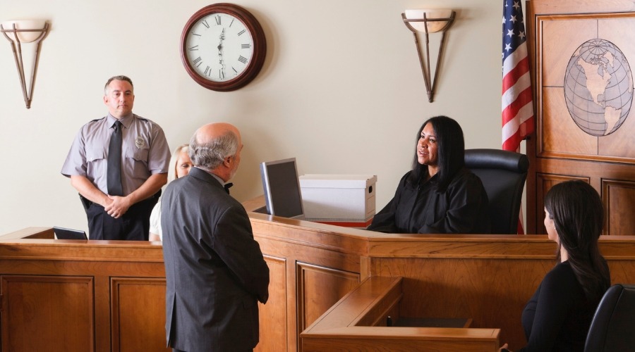 Male attorney speaking to female judge with female client looking to sell structured settlement payments sitting and Bailiff standing in background in courtroom setting with US flag.
